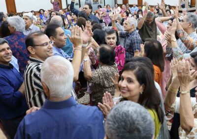 A lively crowd of the Sindhi community celebrating Raksha Bandhan at Gandhi Seva Loka, with men and women dancing, clapping.